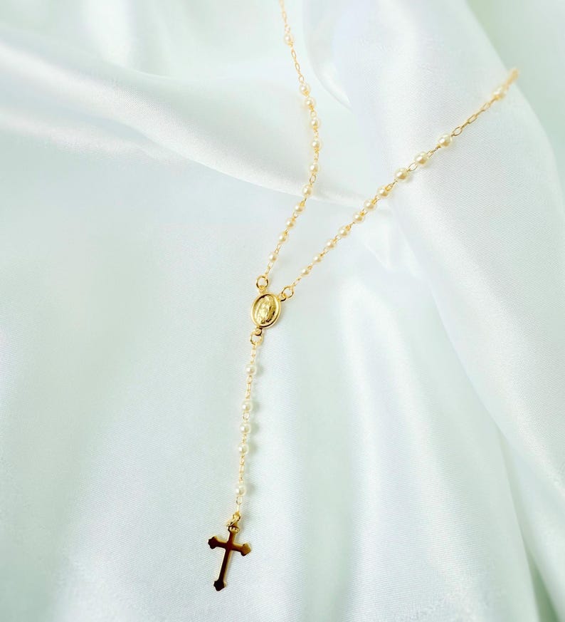 Peut inclure: Un collier de chapelet dor&eacute; avec un pendentif en forme de croix et un petit pendentif ovale repr&eacute;sentant une figure religieuse. Le collier est compos&eacute; de petites perles rondes et est pr&eacute;sent&eacute; sur un tissu blanc et soyeux.