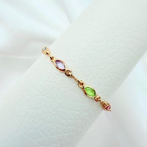 May include: A delicate gold ring with a series of colourful gemstones. The ring features an alternating pattern of oval-shaped purple and green stones, with smaller pink and clear stones. The ring is displayed against a white background.