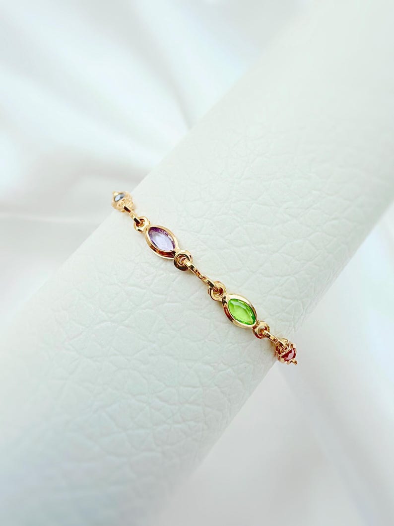 May include: A delicate gold ring with a series of colourful gemstones. The ring features an alternating pattern of oval-shaped purple and green stones, with smaller pink and clear stones. The ring is displayed against a white background.