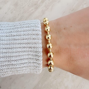 May include: Gold beaded bracelet. The bracelet features a series of round, gold-colored beads strung together. The beads are evenly spaced and have a polished finish. The bracelet is shown on a wrist, with a gray sweater in the background.