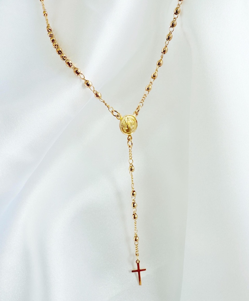 Peut inclure: Collier de chapelet dor&eacute; avec un pendentif en forme de croix. Le collier est compos&eacute; de petites perles rondes et d'un m&eacute;daillon circulaire. La croix et le m&eacute;daillon sont &eacute;galement dor&eacute;s. Le collier est pr&eacute;sent&eacute; sur un fond blanc.