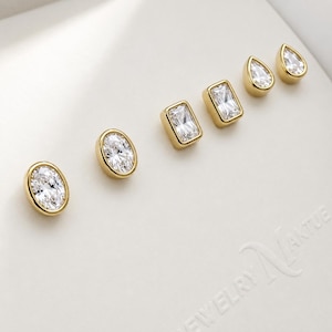 May include: Six gold-toned stud earrings with clear, sparkling cubic zirconia stones. The earrings are in three different shapes: oval, rectangular, and teardrop. The earrings are displayed on a white surface.