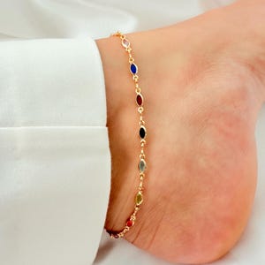May include: Gold-coloured anklet with a chain of colourful, oval gemstones. The gemstones are red, yellow, green, grey, black, and blue. The anklet is worn on an ankle, with a white shirt in the background.
