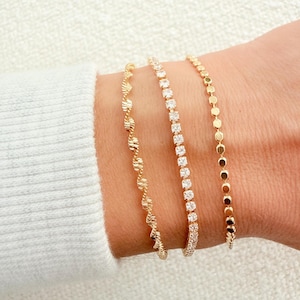 May include: Three gold bracelets are displayed on a wrist. The bracelets include a twisted rope design, a line of square-cut clear stones, and a chain of small, round discs. The bracelets are gold in color.
