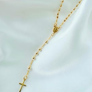 Peut inclure: Collier de chapelet dor&eacute; avec un pendentif en forme de croix. Le collier est compos&eacute; de petites perles rondes et d'un m&eacute;daillon central. La croix est de conception simple. Le collier est pr&eacute;sent&eacute; sur un fond blanc et soyeux.