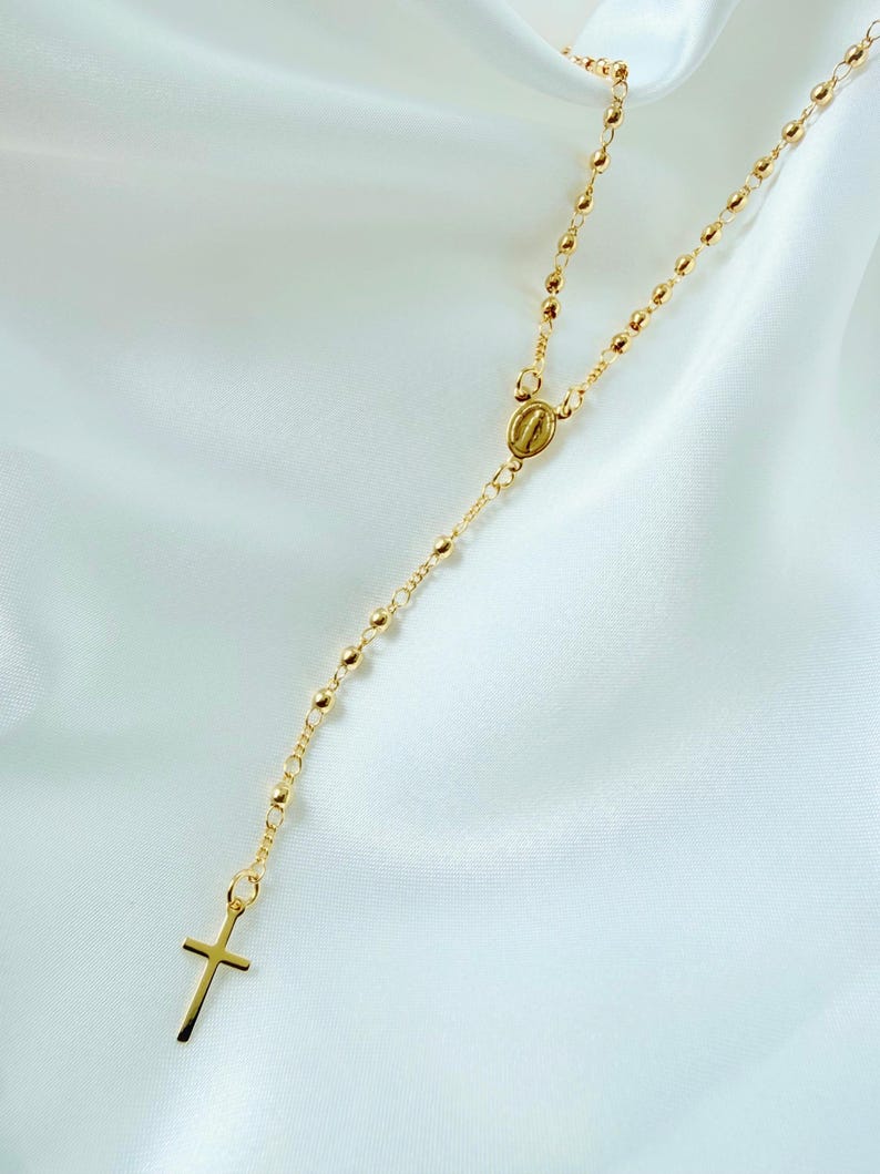 Peut inclure: Collier de chapelet dor&eacute; avec un pendentif en forme de croix. Le collier est compos&eacute; de petites perles rondes et d'un m&eacute;daillon central. La croix est de conception simple. Le collier est pr&eacute;sent&eacute; sur un fond blanc et soyeux.