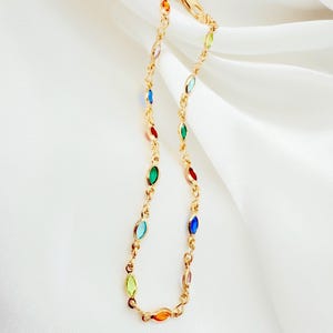 May include: A gold necklace with a chain design, featuring colourful, oval-shaped gemstones in various colours. The necklace is displayed against a white, draped fabric background.
