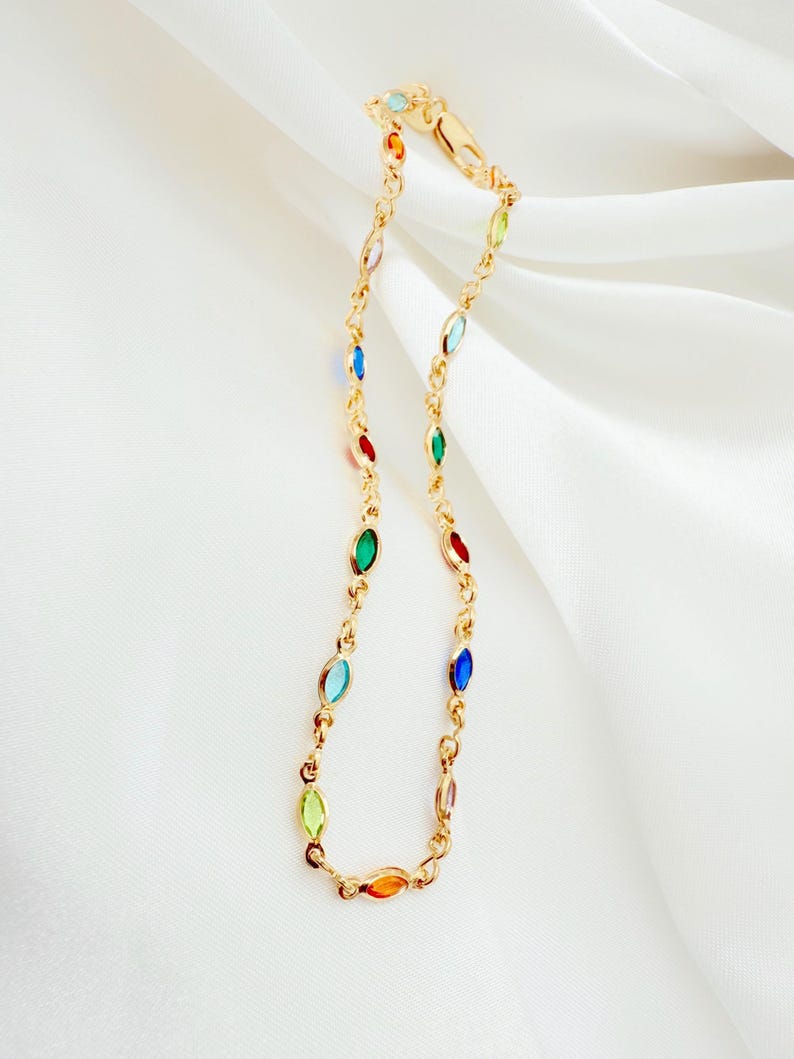 May include: A gold necklace with a chain design, featuring colourful, oval-shaped gemstones in various colours. The necklace is displayed against a white, draped fabric background.