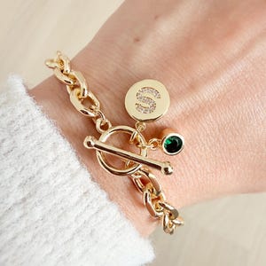 Könnte beinhalten: Goldfarbenes Armband mit Knebelverschluss. Das Armband hat ein Kettenglied-Design und einen runden Anhänger mit dem Buchstaben "S", der mit kleinen Kristallen besetzt ist. Ein kleiner grüner Edelstein hängt am Anhänger.