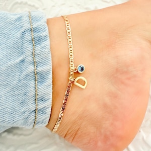 May include: Gold-colored anklet with a chain link design. The anklet features a small blue gemstone and a gold letter "D" charm. The anklet is worn on an ankle, with a denim jean visible in the background.