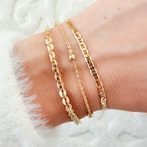 May include: Three gold-colored bracelets of varying designs are displayed on a wrist. The bracelets include a chain link style, a delicate chain with a small bead, and a bracelet with a series of connected circular shapes. The bracelets are set against a white background.