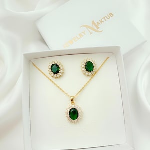 May include: A gold-coloured jewellery set comprising a necklace and earrings, presented in a white box. Each piece features a large, oval, emerald-green gemstone encircled by smaller, clear stones. The box lid displays the text "JEWELRY Maktub".