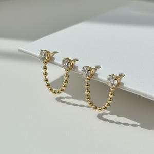 May include: A pair of gold-colored double-pierced earrings. Each earring features a small, clear gemstone at the top and a chain of small, gold-colored beads that dangles below. The earrings are displayed on a white surface.