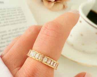 May include: A gold-toned ring with rectangular, clear gemstones set in a row. The ring is on a person's finger, with a soft pink nail polish. The background includes a blurred flower and a cup.