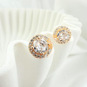 May include: A pair of gold-toned stud earrings with a halo of small, clear crystals surrounding a larger, clear, round-cut crystal. The earrings are displayed on a white, textured surface, creating a contrast with the sparkling jewelry.