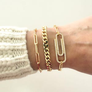 Könnte beinhalten: Drei goldene Armbänder werden gezeigt. Das erste ist ein filigranes Kettenglied-Design, das zweite eine dickere Panzerkette und das dritte ein Armband im Büroklammer-Stil, das mit kleinen, funkelnden Steinen besetzt ist. Die Armbänder werden an einem Handgelenk getragen.