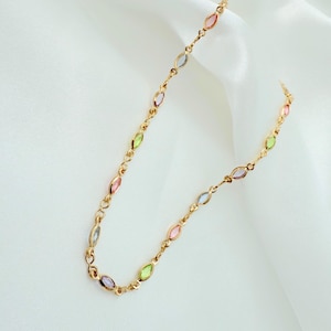 May include: A gold-coloured necklace with a chain and a series of colourful, oval-shaped gemstones. The gemstones are in shades of pink, green, blue, and purple. The necklace is displayed on a white, textured background.