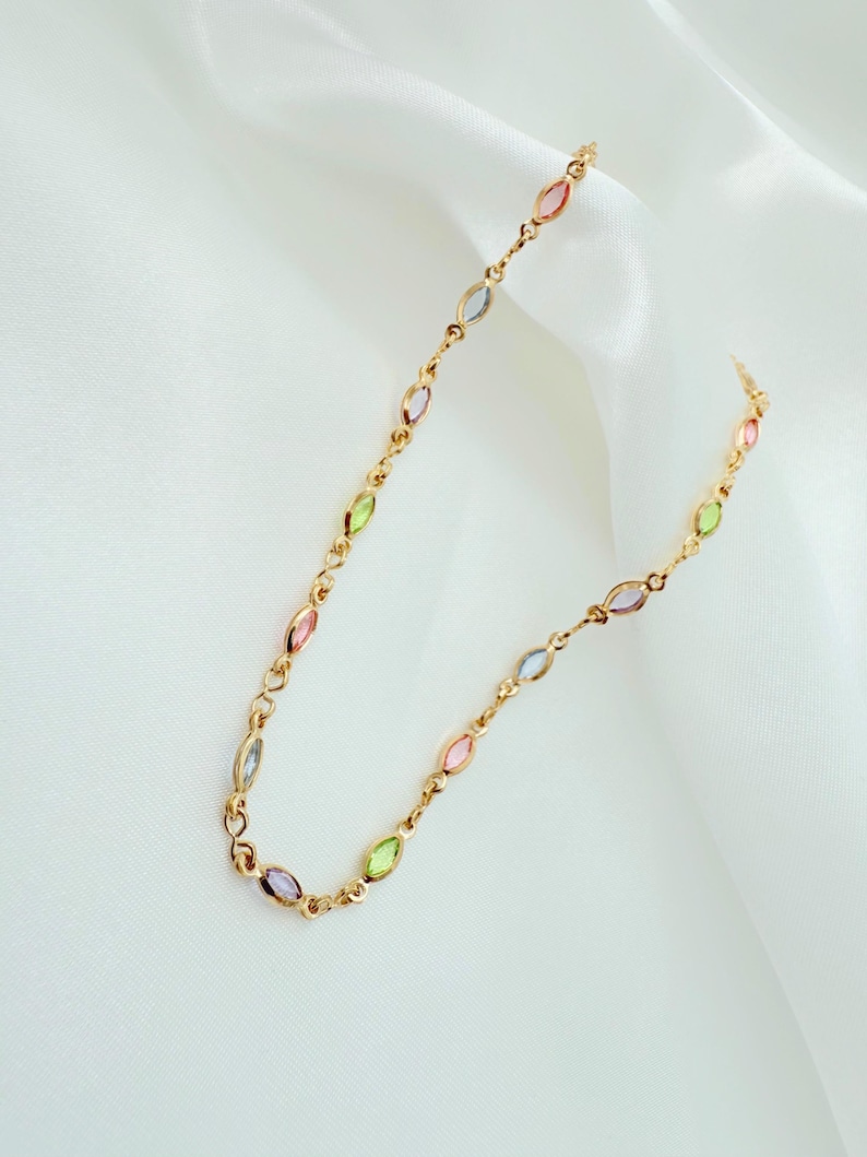 May include: A gold-coloured necklace with a chain and a series of colourful, oval-shaped gemstones. The gemstones are in shades of pink, green, blue, and purple. The necklace is displayed on a white, textured background.
