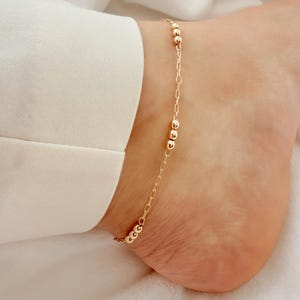 May include: Gold-colored anklet featuring a delicate chain interspersed with small, round beads. The anklet is worn on an ankle, with a white shirt visible in the background. The jewelry is simple and elegant.