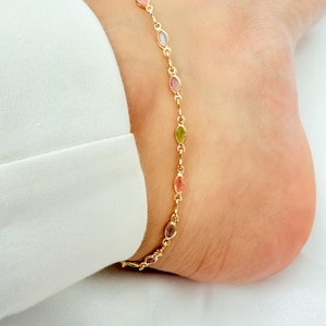 May include: A gold-coloured anklet featuring oval-shaped, multi-coloured gemstones. The gemstones are in shades of pink, purple, green, and yellow. The anklet is worn on an ankle.