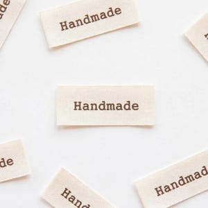 Könnte beinhalten: Mehrere cremefarbene Stoffetiketten mit dem braun gedruckten Wort "Handmade". Die rechteckigen Etiketten sind auf einem weißen Hintergrund verstreut. Der Text ist auf jedem Etikett zentriert.