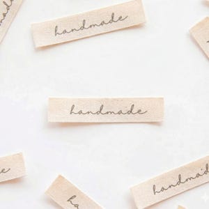May include: Several small, rectangular fabric labels with the word "handmade" printed in a cursive font. The labels are a light beige color and are scattered across a white surface.