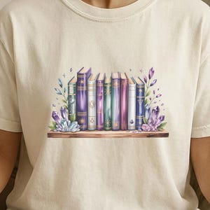 Può includere: Una t-shirt a maniche corte color avorio con una stampa grafica di una fila di libri colorati su uno scaffale in legno, arricchita da grappoli di cristalli viola e blu e piccoli rametti floreali alle estremità della collezione.