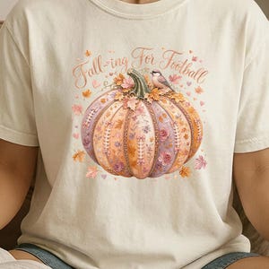 May include: An ivory short-sleeve t-shirt featuring a graphic of a pumpkin decorated with floral patterns, autumn leaves, and a small bird. The text "Fall-ing For Football" is printed in a cursive font above the pumpkin illustration.