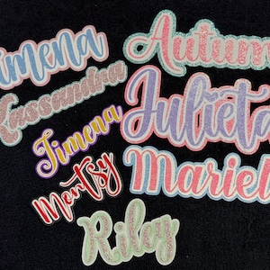May include: A collection of custom name cutouts featuring glittery finishes in various colors including pink, blue, purple, yellow, and red. The names Jimena, Kassandra, Julieta, Mariel, Montsy, Autumn, and Riley are written in a cursive script font.