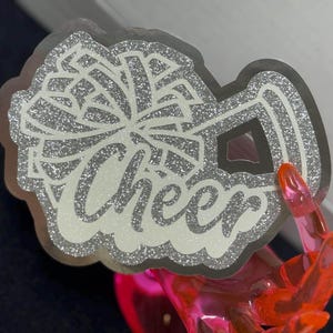 May include: A silver glitter sticker featuring a stylized graphic of a pom-pom and a megaphone with the word Cheer written in a cursive script font. The design has a clear border and a textured, sparkling finish throughout the entire surface.