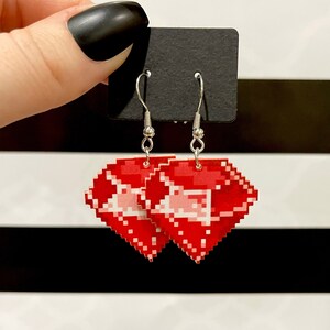 May include: A pair of silver dangle earrings with red pixelated diamond-shaped charms. The earrings are hanging from a black card.