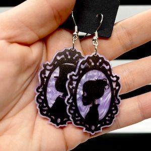 May include: A pair of purple and black earrings featuring a silhouette of a couple in a frame. The earrings are made of acrylic and have silver hooks.