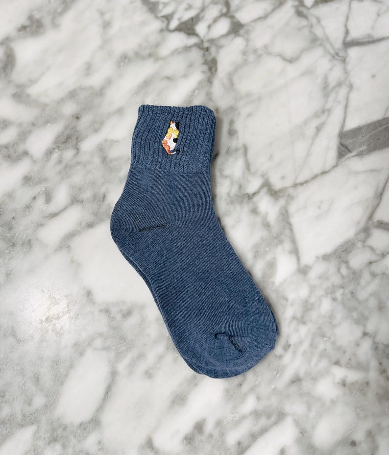 May include: A pair of blue ankle socks with a colorful cat embroidered on the top of the sock.
