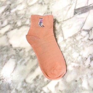 May include: A pair of peach-colored ankle socks with a small embroidered cat design on the front.