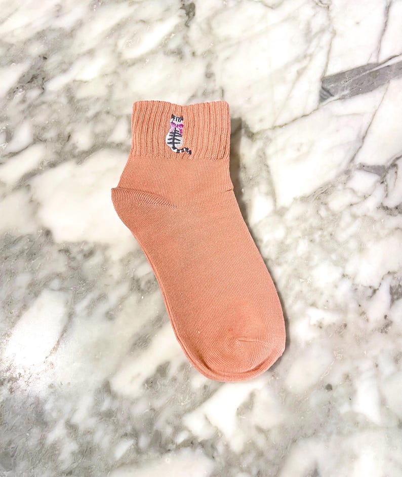 May include: A pair of peach-colored ankle socks with a small embroidered cat design on the front.