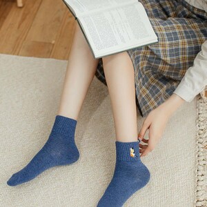 May include: A pair of blue ankle socks with a small embroidered design on one sock. The socks are worn by a person sitting on a rug.