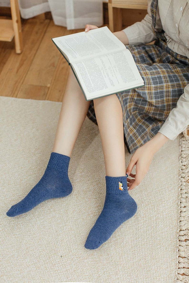 May include: A pair of blue ankle socks with a small embroidered design on one sock. The socks are worn by a person sitting on a rug.