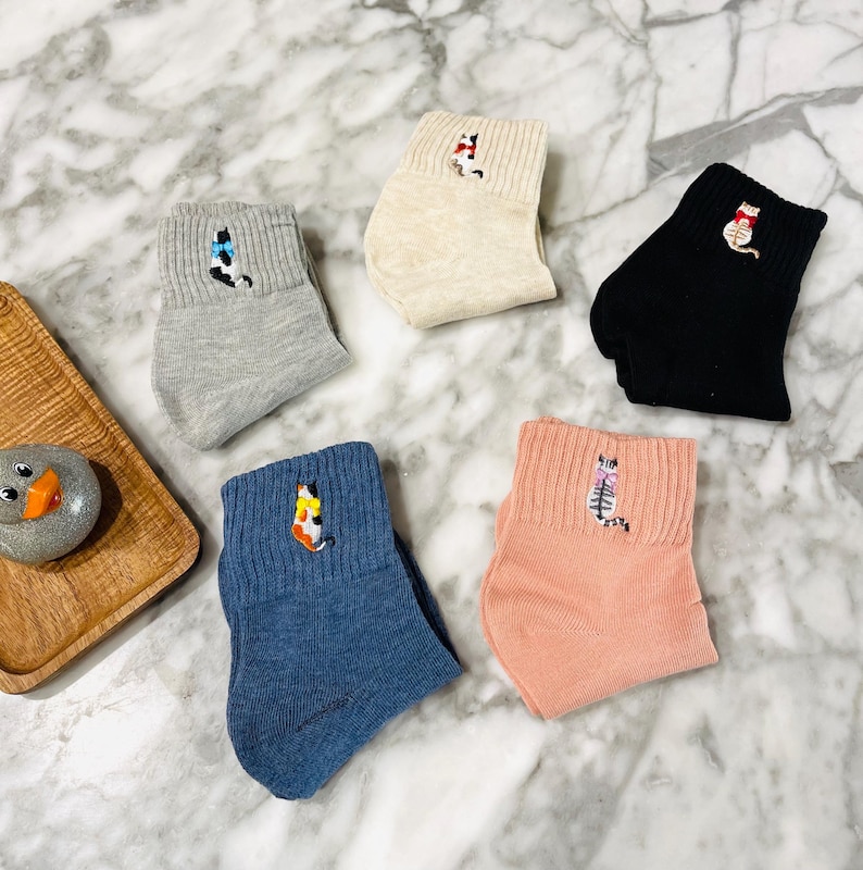May include: Six pairs of ankle socks with embroidered cat designs. The socks are in various colors, including gray, beige, black, blue, and pink.