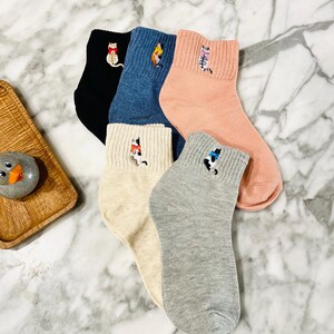 May include: Five pairs of ankle socks with embroidered cat designs. The socks are in various colors, including black, blue, pink, beige, and gray.