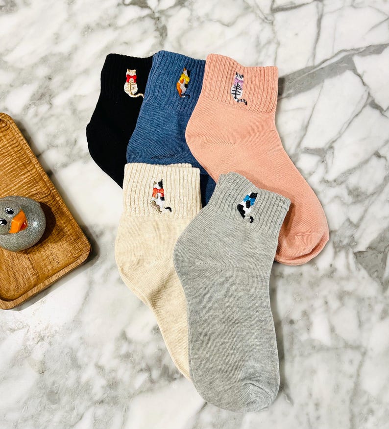 May include: Five pairs of ankle socks with embroidered cat designs. The socks are in various colors, including black, blue, pink, beige, and gray.