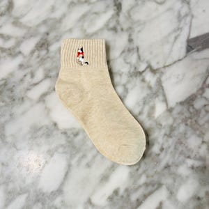 May include: A pair of beige ankle socks with an embroidered cat design. The cat is wearing a red scarf.