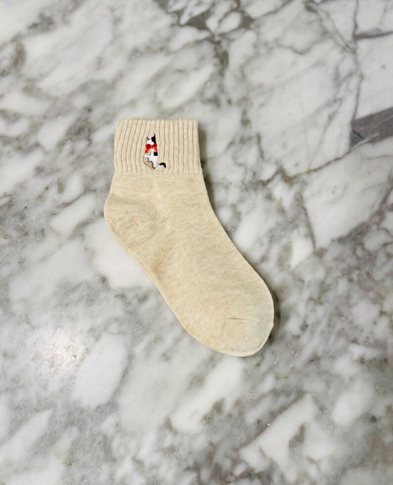 May include: A pair of beige ankle socks with an embroidered cat design. The cat is wearing a red scarf.