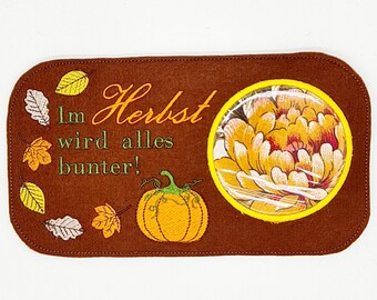 May include: Brown felt rectangle with embroidered text "Im Herbst wird alles bunter!" and a pumpkin, a flower, and leaves.