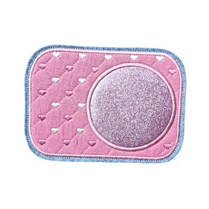 May include: A pink rectangular patch with a quilted heart pattern and a glittery circle. The patch has a light blue border. The hearts are white. This patch is likely for decorative purposes.