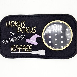 May include: Black felt coaster with a yellow stitched border. The coaster features a witch's hat, a broom, and a circle with white polka dots. The text "Hokus Pokus 3x schwarzer Kaffee" is embroidered in yellow.