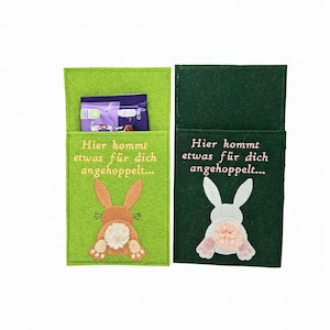 Könnte beinhalten: Zwei Oster-Filztaschen, eine grüne und eine dunkelgrüne. Die grüne Tasche hat ein braunes Hasenmuster und den Text "Hier kommt etwas für dich angehoppelt...". Die dunkelgrüne Tasche hat ein weißes Hasenmuster und denselben Text.
