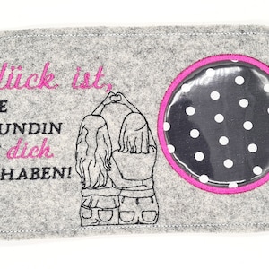 Könnte beinhalten: Grauer Filzstoff mit rosa Nähten und einem schwarzen Kreis mit weißen Punkten und einer durchsichtigen Plastikabdeckung. Der Text "Glück ist, eine Freundin wie dich zu haben!" ist in rosa gestickt.