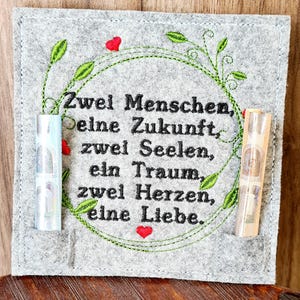 Op de afbeelding: Een grijze wollen wandhanger met geborduurde groene wijnstokken en rode harten. De tekst luidt "Zwei Menschen, Deine Zukunft, zwei Seelen, ein Traum, zwei Herzen, eine Liebe."