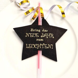 Può includere: Decorazione a forma di stella nera con testo dorato che recita "Bring das NEUE JAHR zum LEUCHTEN!". Una cannuccia rosa e trasparente è inserita nella stella. Nastri dorati e bianchi sono sullo sfondo. La decorazione è su un piatto bianco.