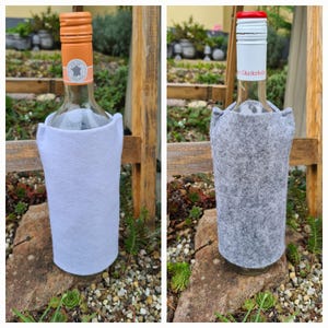 Pode incluir: Duas garrafas de vinho com capas de feltro para garrafas. A garrafa à esquerda tem uma capa branca e a garrafa à direita tem uma capa cinzenta. A garrafa à esquerda tem um rótulo que diz "In the Country". A garrafa à direita tem um rótulo que diz "Glücksfee".